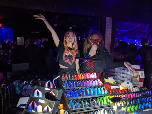 Harley and Irene stand behind a table with many colorful devil horns for sale. Irene has a butterfly emoji covering her face.