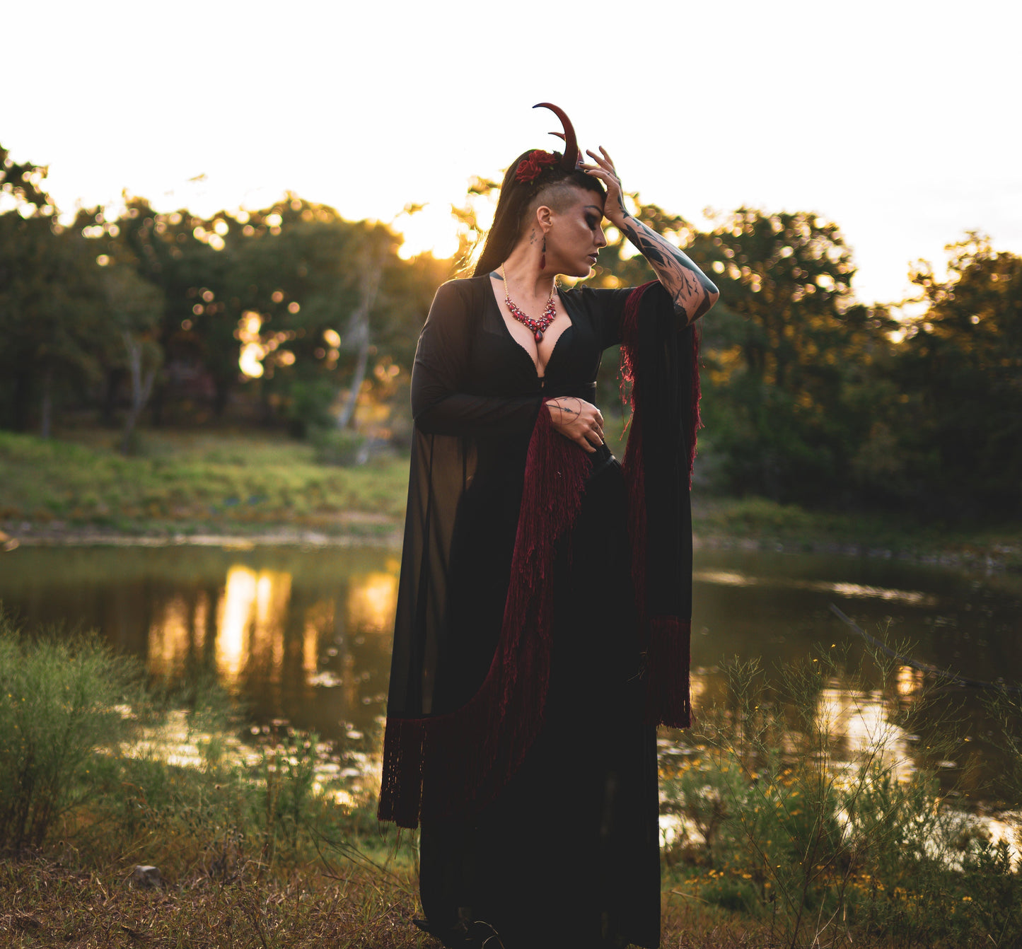 Zoe Sol models Azazel horns by Haus of Brimstone in a black and red witchy ensemble, their hand to their head, in a field at golden hour. Photo by Optic Onslaught.