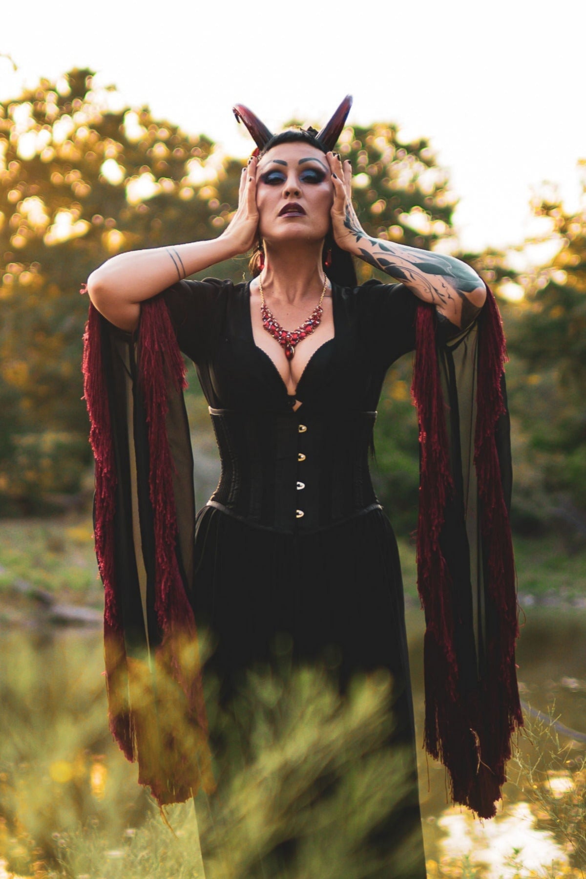 Zoe Sol models Azazel horns by Haus of Brimstone in a black and red witchy ensemble, their hands at the side of their face, in a field at golden hour. Photo by Optic Onslaught.