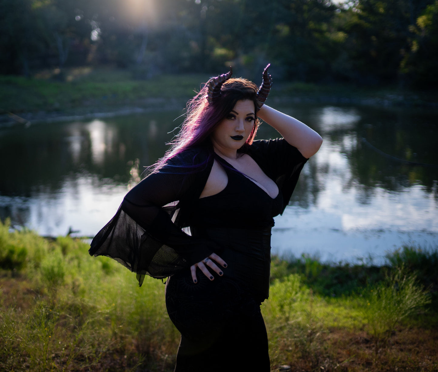 Lady Marionette models Tiefling horns by Haus of Brimstone in a black witchy ensemble, in front of a pond in a field at golden hour. Photo by Optic Onslaught.