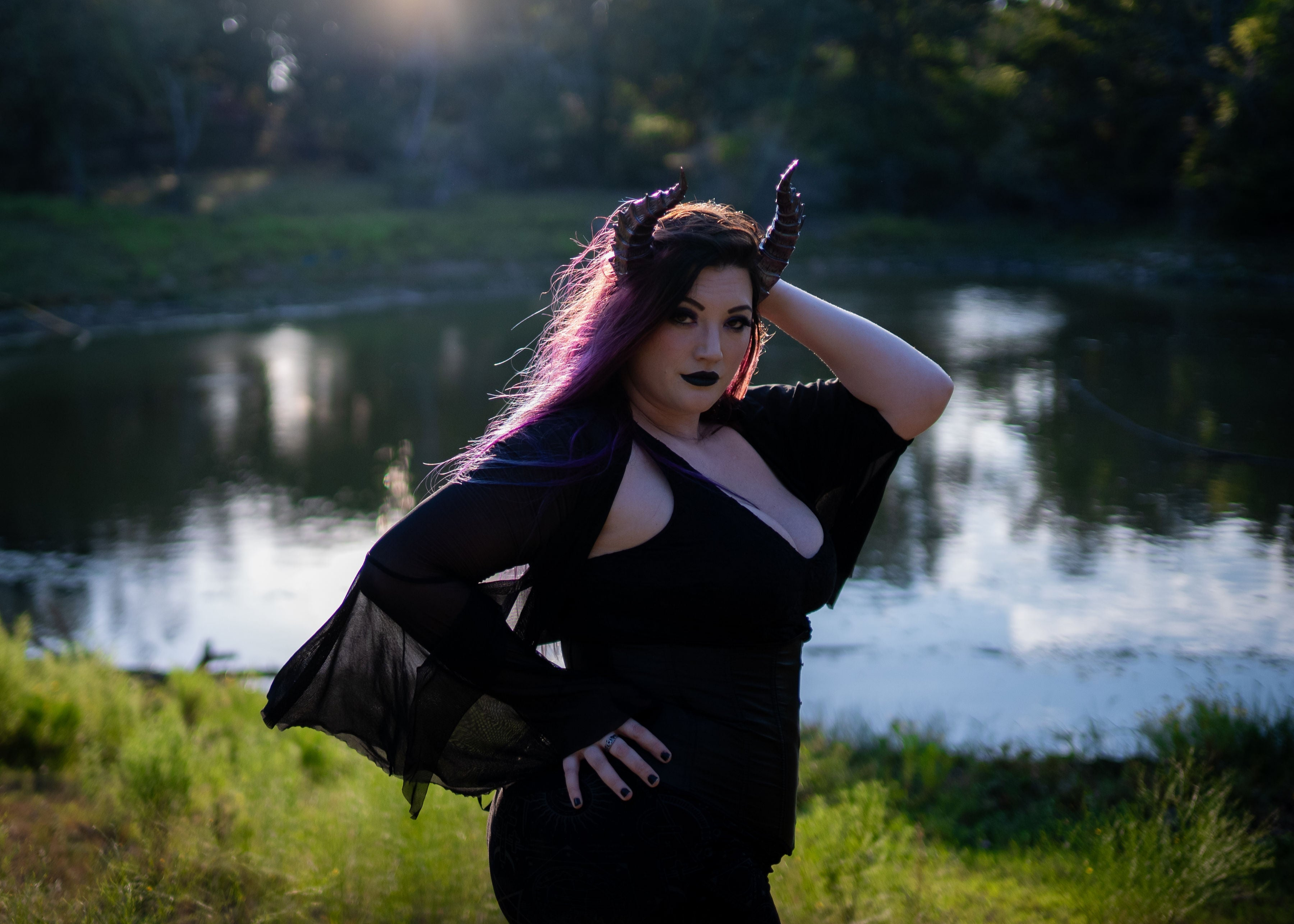 Lady Marionette models Tiefling horns by Haus of Brimstone in a black witchy ensemble, in front of a pond in a field at golden hour. Photo by Optic Onslaught.
