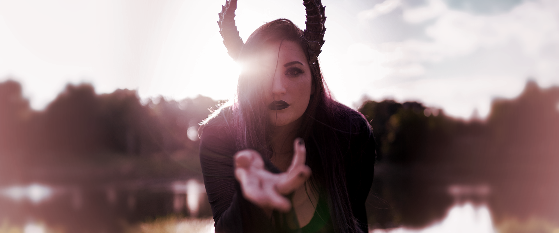 Lady Marionette models Tiefling horns by Haus of Brimstone in a black witchy ensemble, in front of a pond in a field at golden hour. Photo by Optic Onslaught.