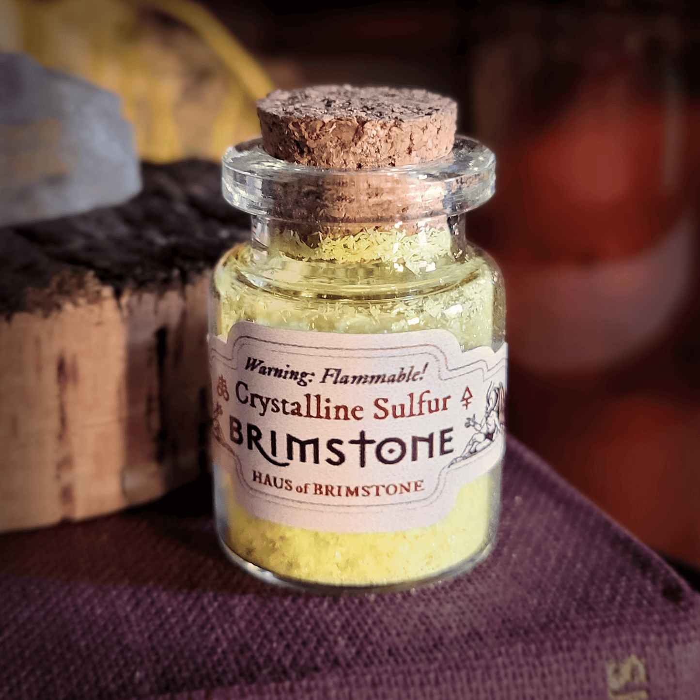 A tiny bottle of crystallized sulfur with a cork stopper sits on an old book. The label reads, “Warning: Flammable! Crystalline Sulfur. Brimstone. Haus of Brimstone.” The label also depicts two devils and two different alchemical symbols for brimstone.