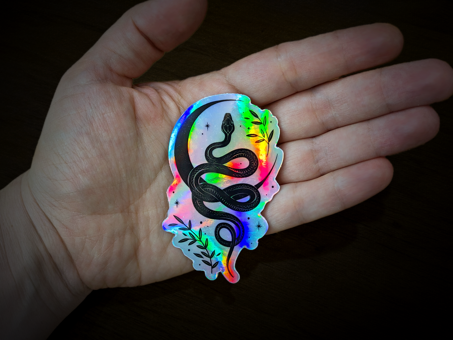 A holographic snake sticker featuring a moon, sparkles, and leaves by artist Croisy.