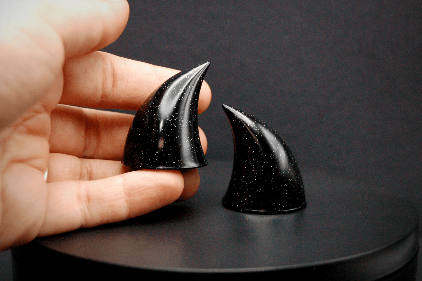 A hand holding one of a pair of small black devil horns with a glitter finish.
