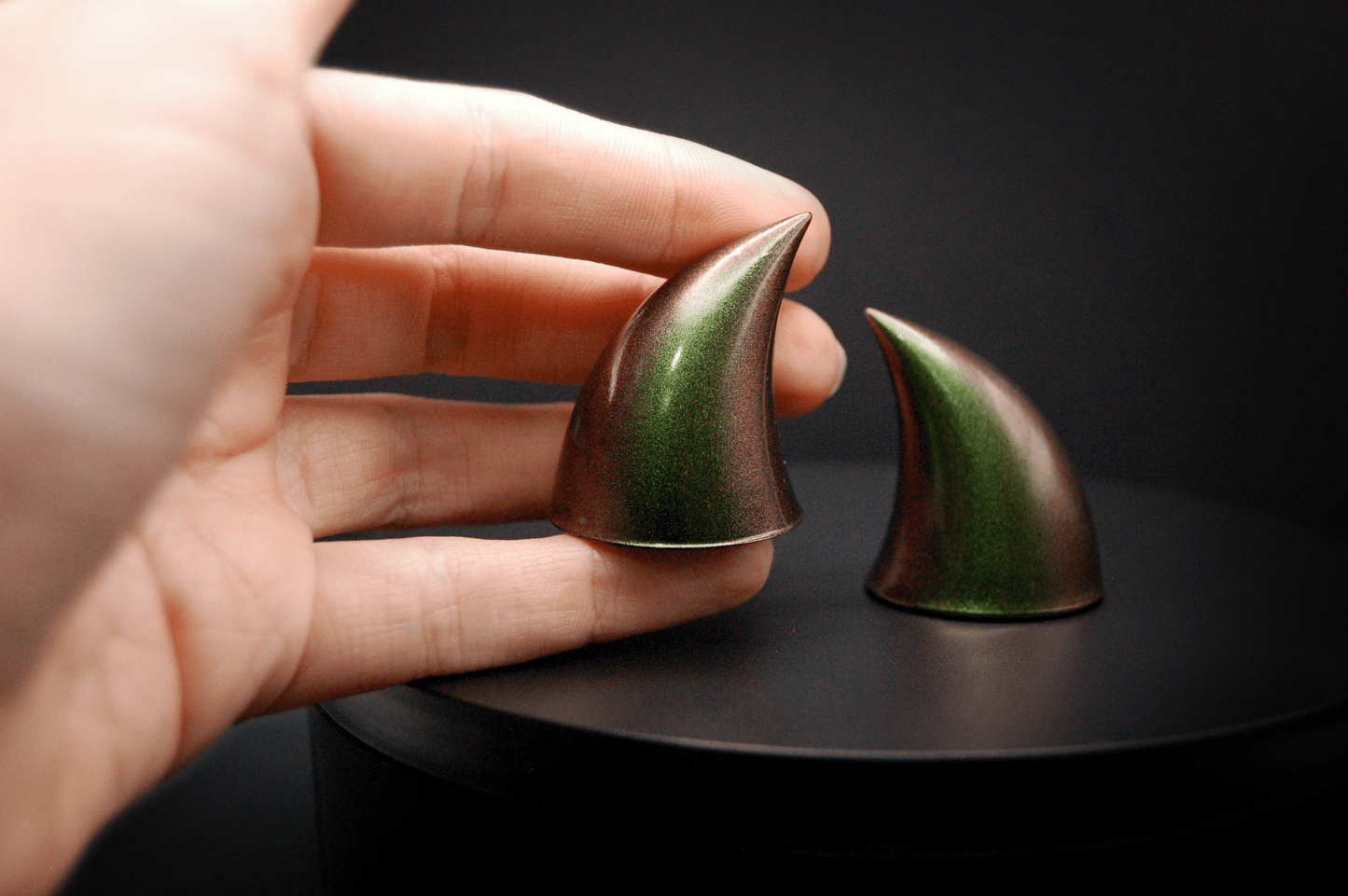 A hand holding an imp devil horn in metallic coppery green.