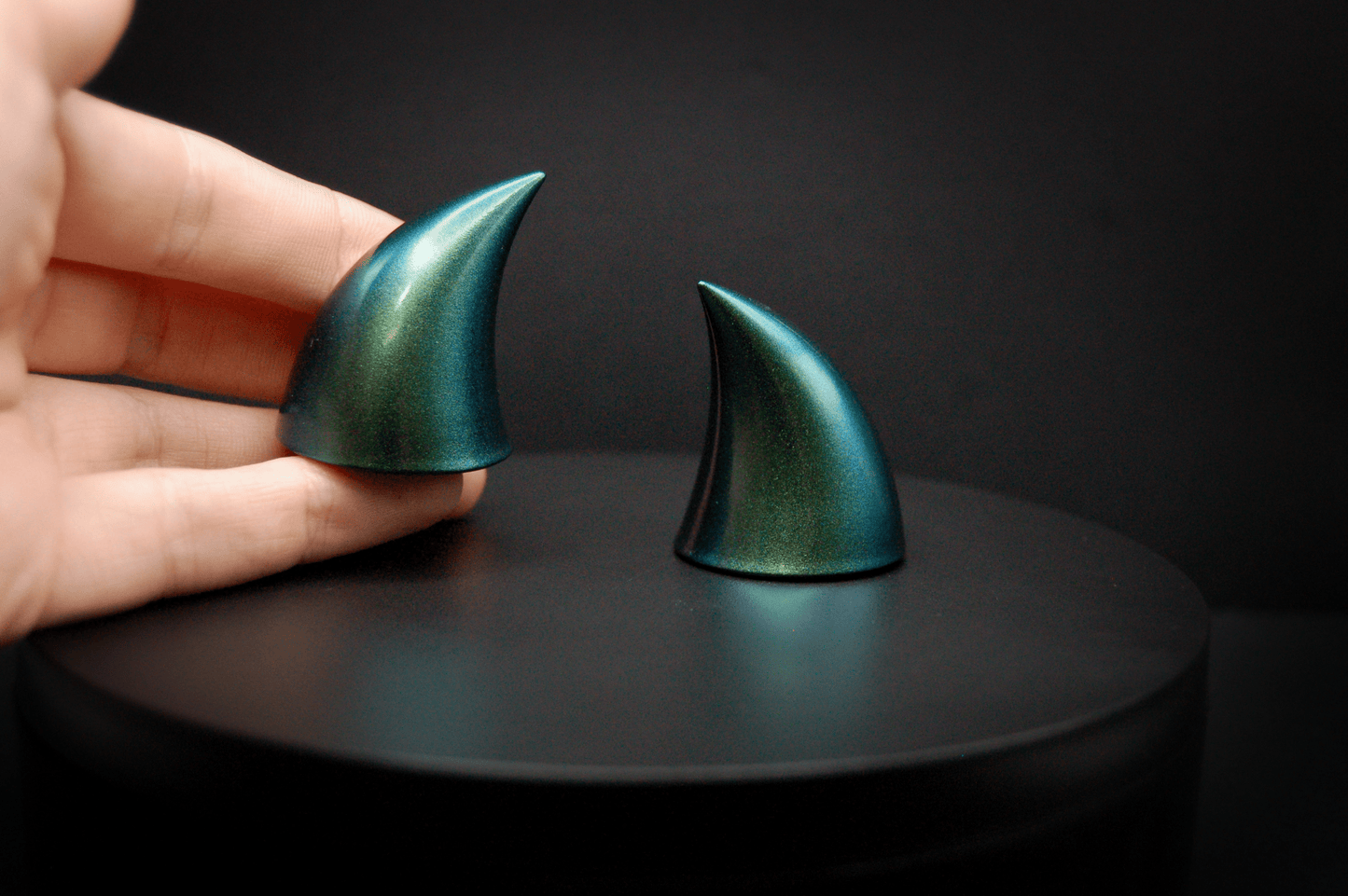 A hand holding an imp devil horn in metallic blue-green.