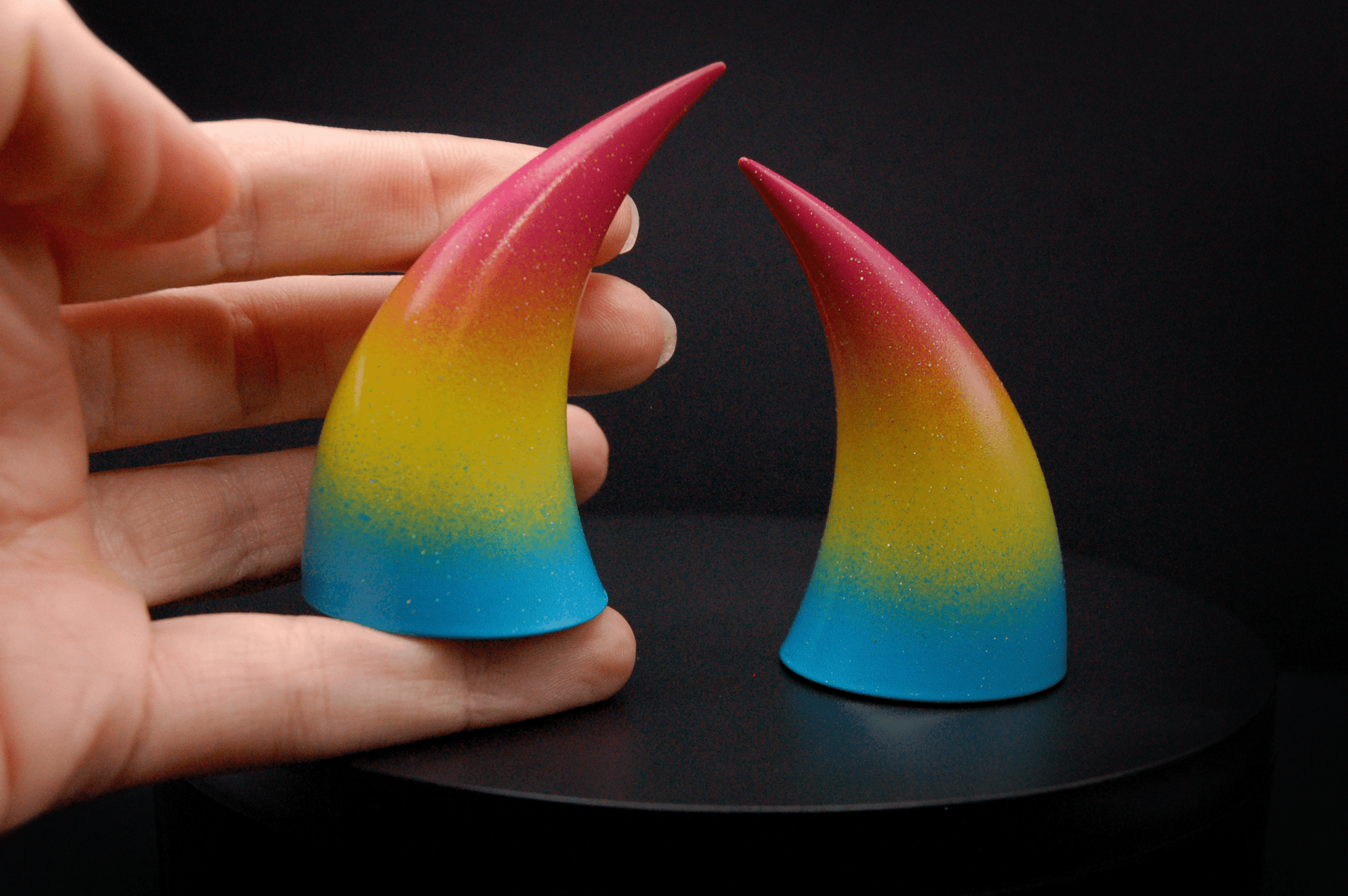 A hand holding an imp devil horn in a  sky blue, to bright yellow, to warm pink ombré, the colors of the pansexual pride flag  with a glittery finish.