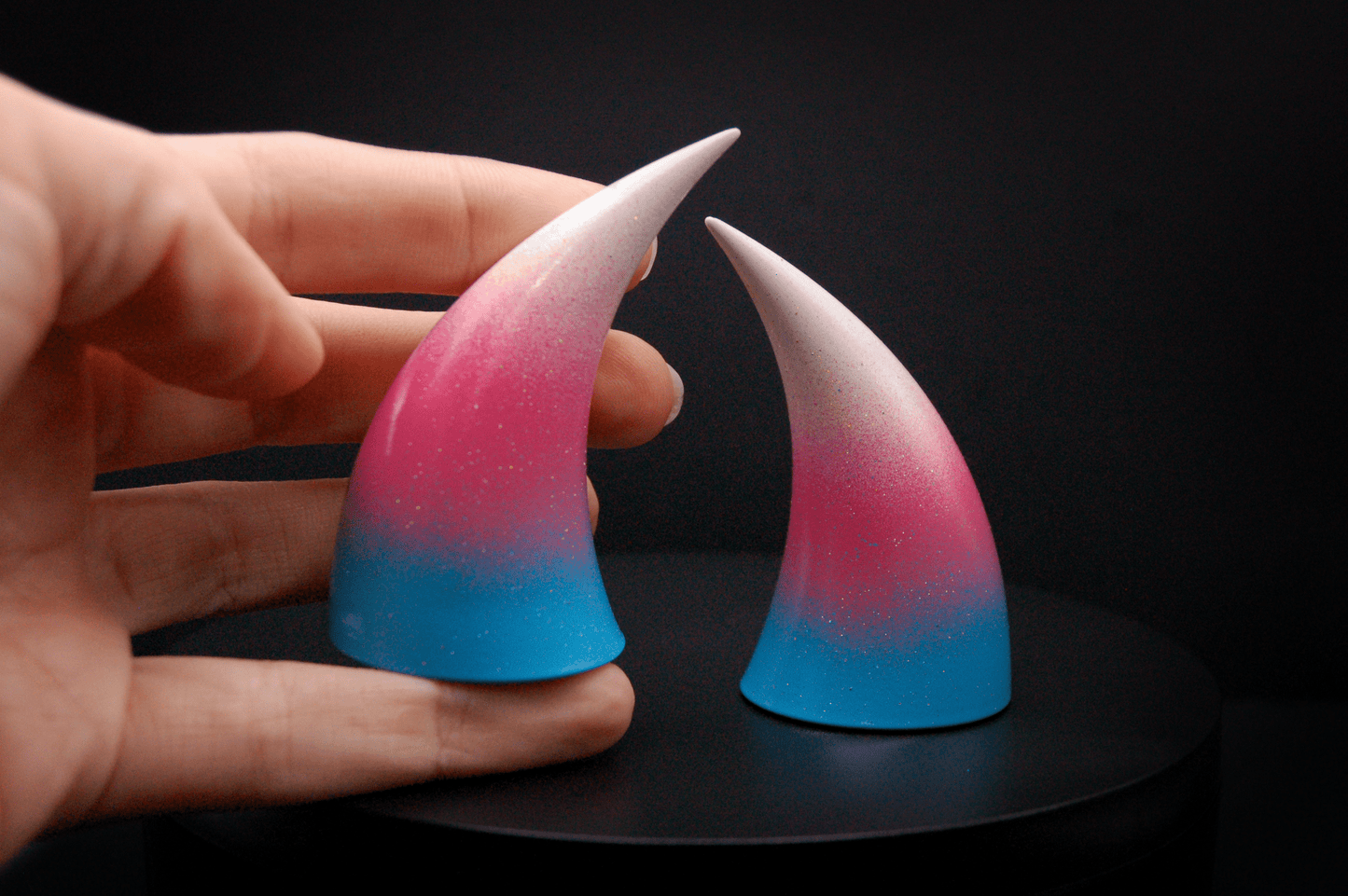A hand holding an imp devil horn in a light blue, medium pink, and white ombré, the colors of the trans pride flag with a glittery finish.