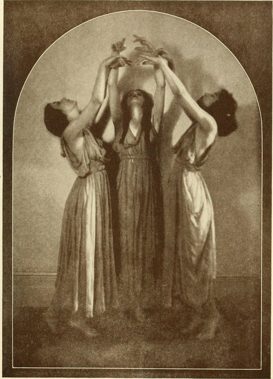 Three women in classical robes facing each other and holding their arms together in the air in a mystical pose.