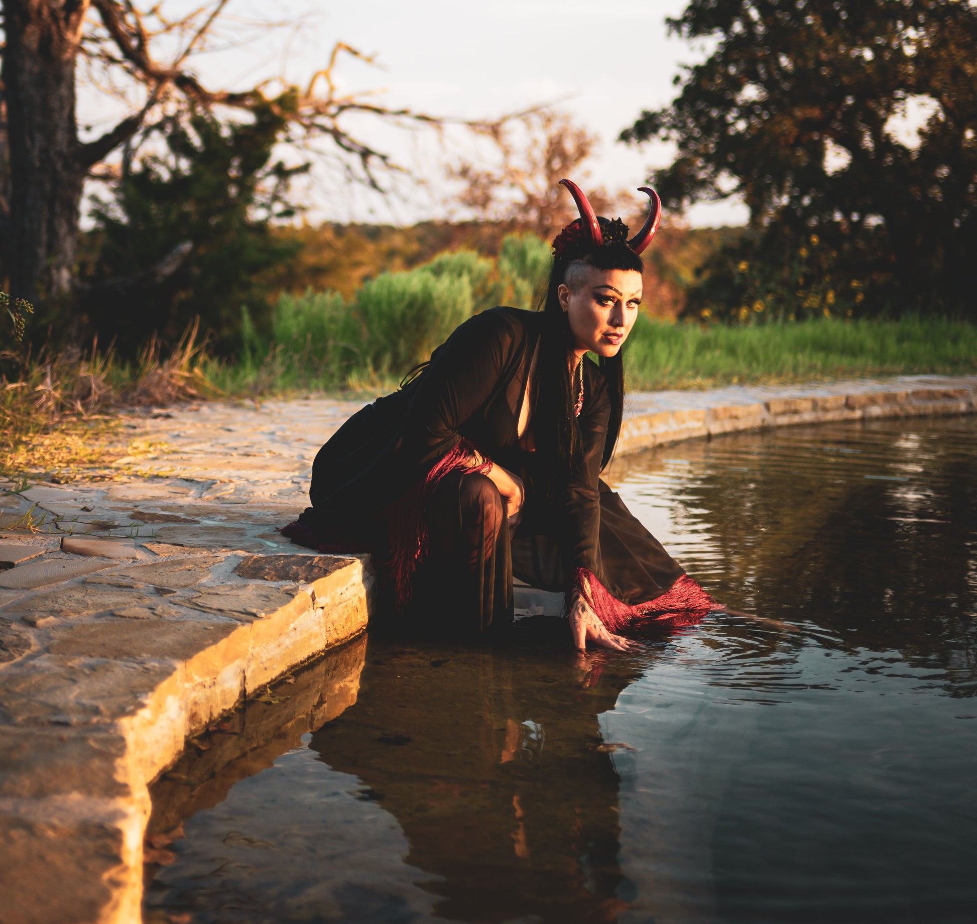 Zoe Sol models Azazel horns by Haus of Brimstone in a black and red witchy ensemble at the edge of a pond, their hand in the water. Photo by Optic Onslaught.