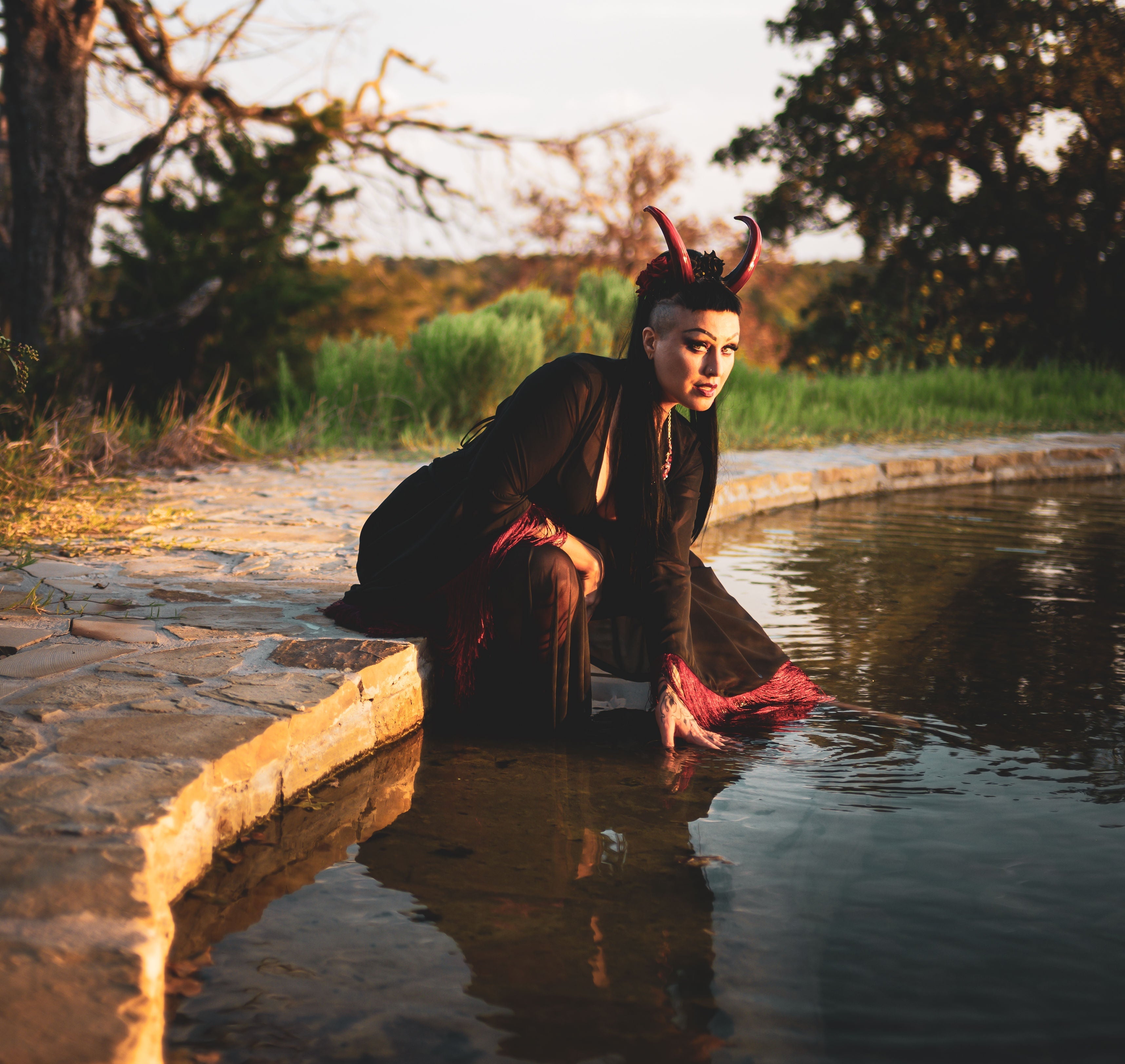 Zoe Sol models Azazel horns by Haus of Brimstone in a black and red witchy ensemble at the edge of a pond, their hand in the water. Photo by Optic Onslaught.