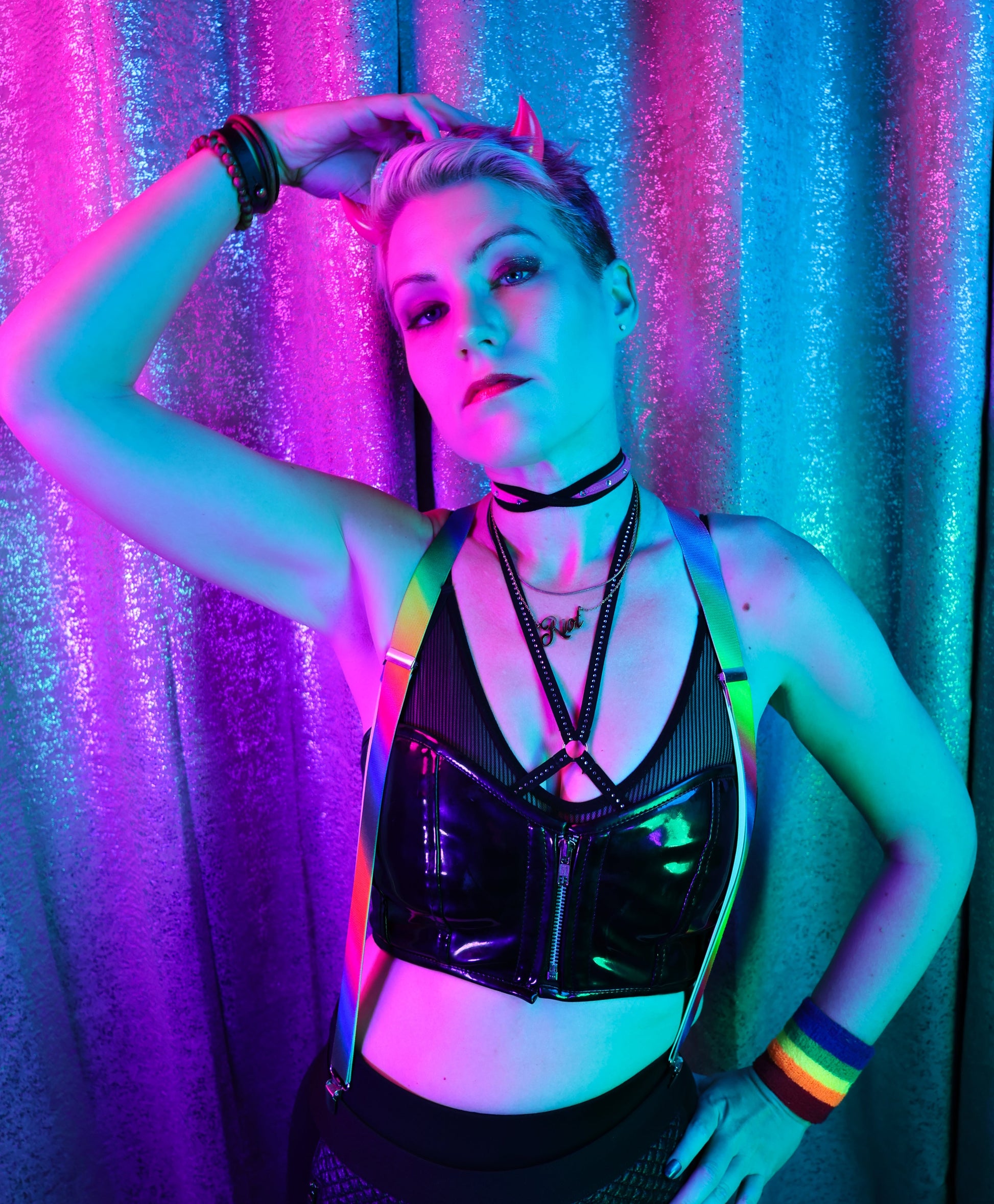 Reignbow Riot wears Hot Pink imp horns with a black outfit and rainbow accessories under neon lights against a shimmery backdrop. Image by Synthetic Studio.