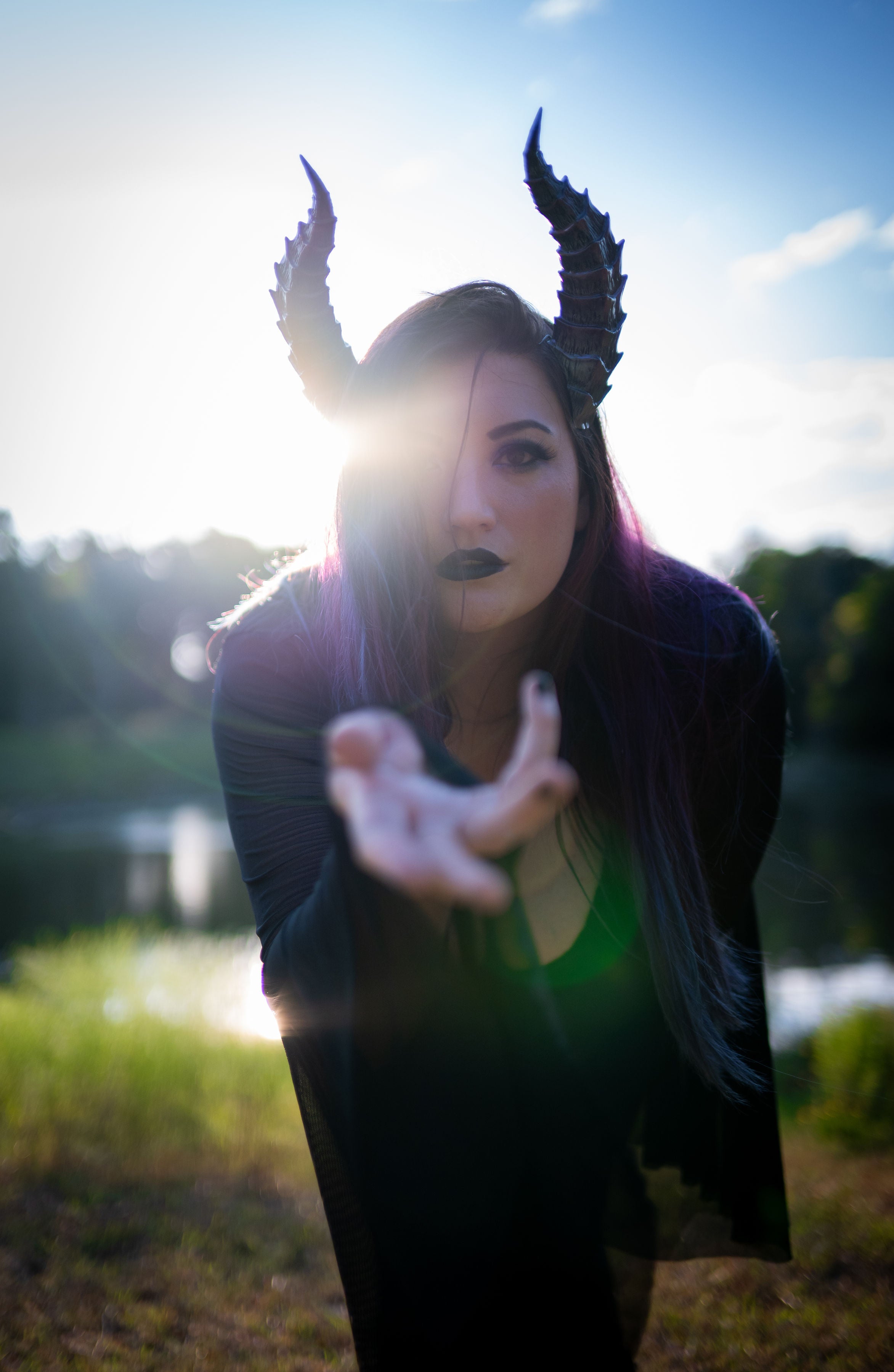 Lady Marionette models Tiefling horns by Haus of Brimstone in a black witchy ensemble, reaching out toward the camera, in a field at golden hour. Photo by Optic Onslaught.