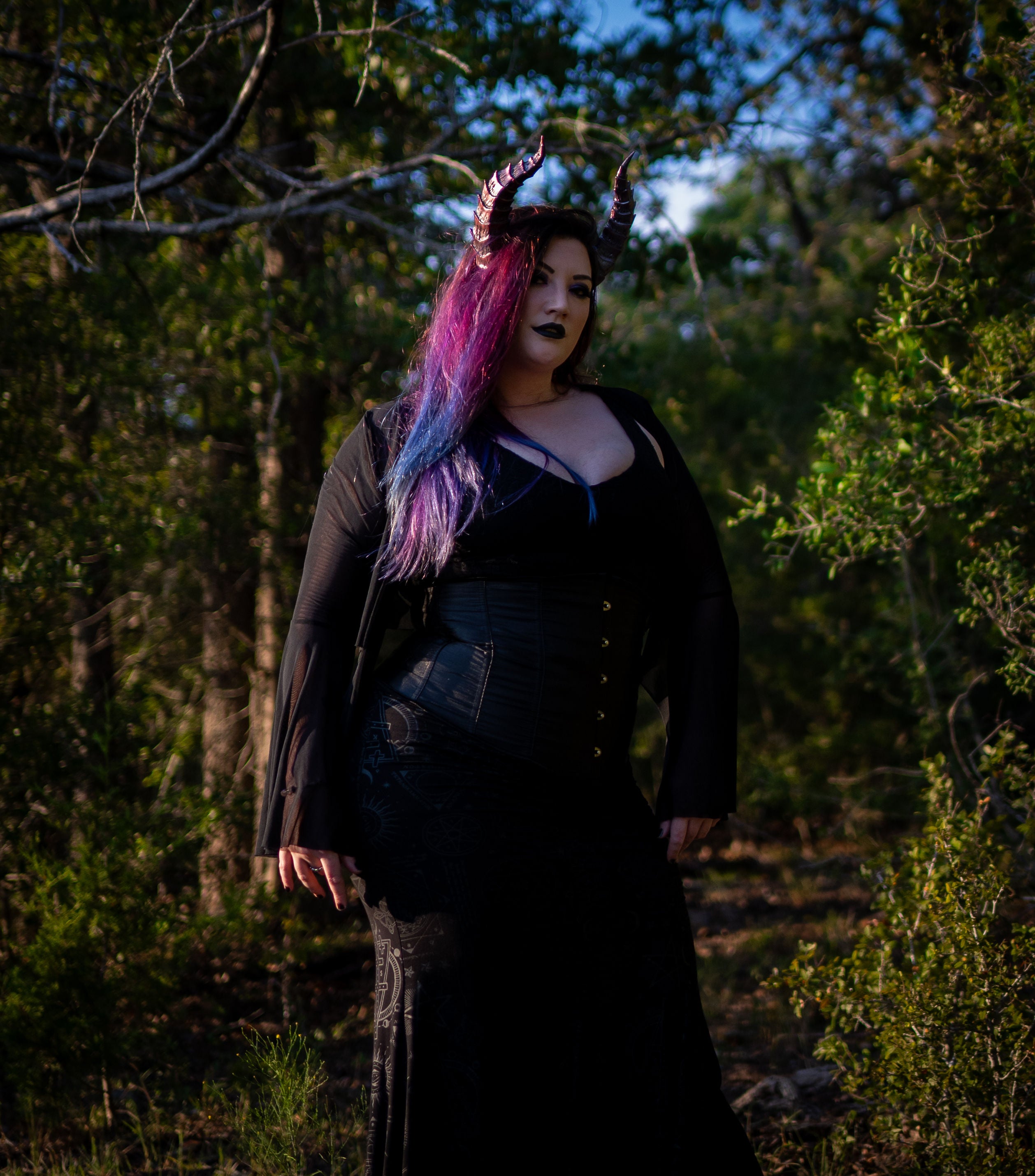 Lady Marionette models Tiefling horns by Haus of Brimstone, wearing a black witchy ensemble, posed in front of a stand of trees. Photo by Optic Onslaught.