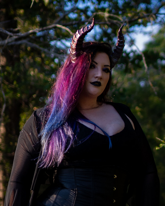 Lady Marionette models Tiefling horns by Haus of Brimstone, wearing a black witchy ensemble, posed in front of a stand of trees. Photo by Optic Onslaught.