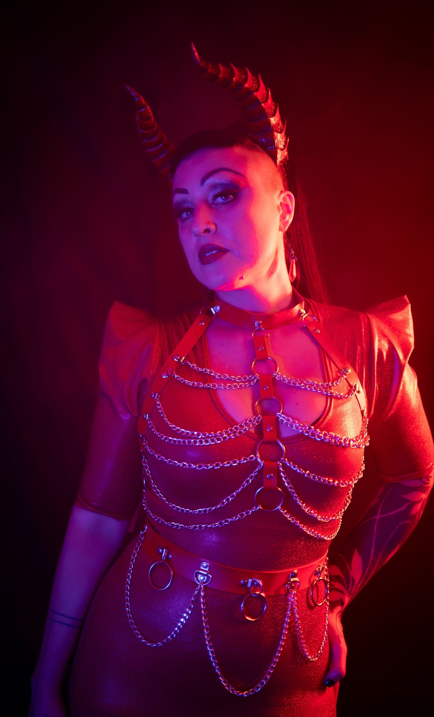Zoe Sol models scarlet Tiefling horns by Haus of Brimstone in a glossy red ensemble with harness and chains. Photo by Optic Onslaught.